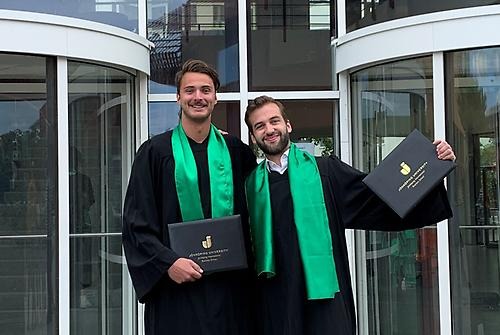 Niklas Koch and Sören Pongratz in their graduation gowns stand outside JIBS entrance. They have their arms around each other's shoulders and have their diplomas raised in the air. Both are smiling and looking out to the camera.